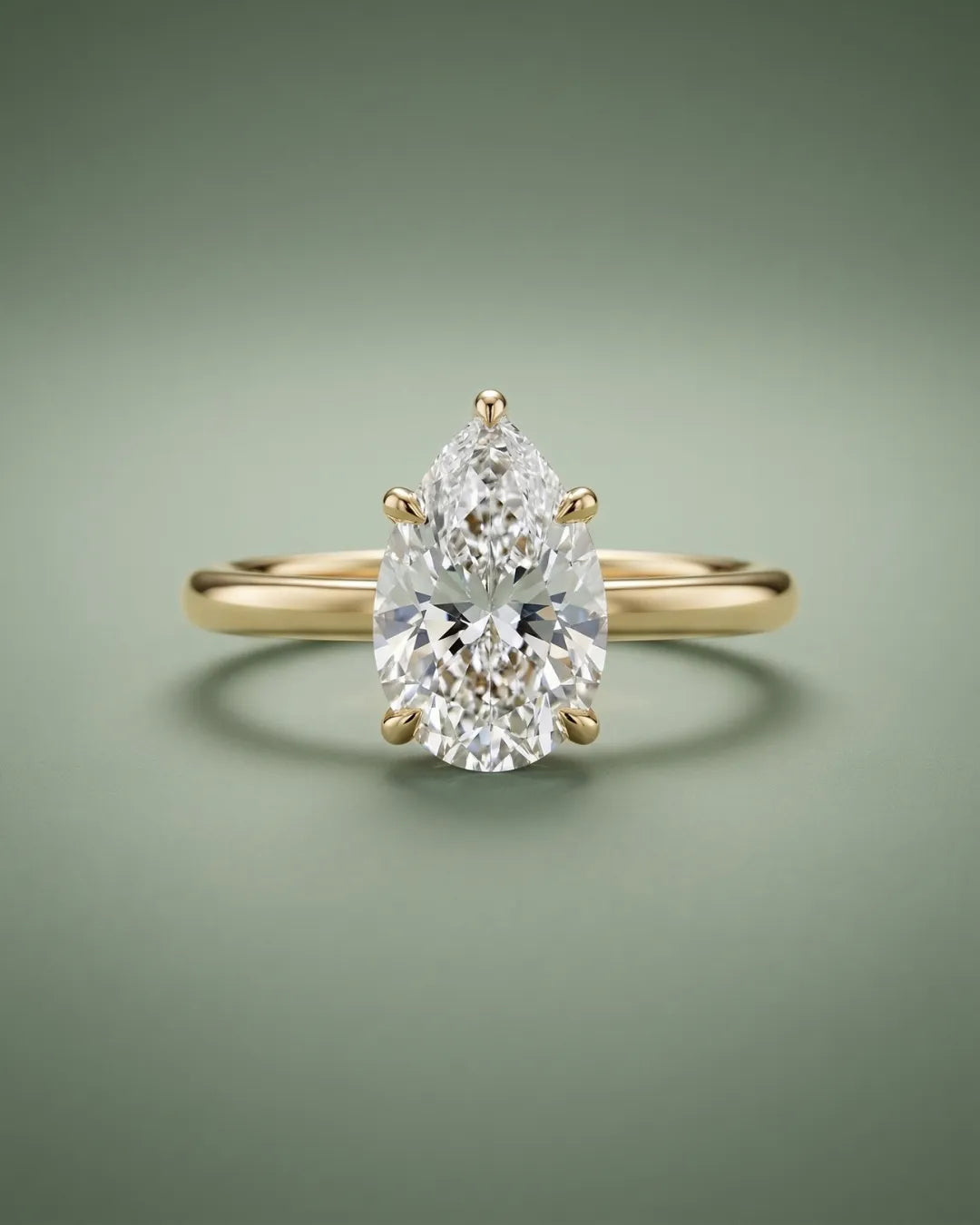 Nærbilde av en pear-slipet lab-diamant forlovelsesring i gult gull med slank 1,8 mm skinne og stor dråpeformet diamant fattet i fem klør, fotografert mot dempet sagegreen bakgrunn.