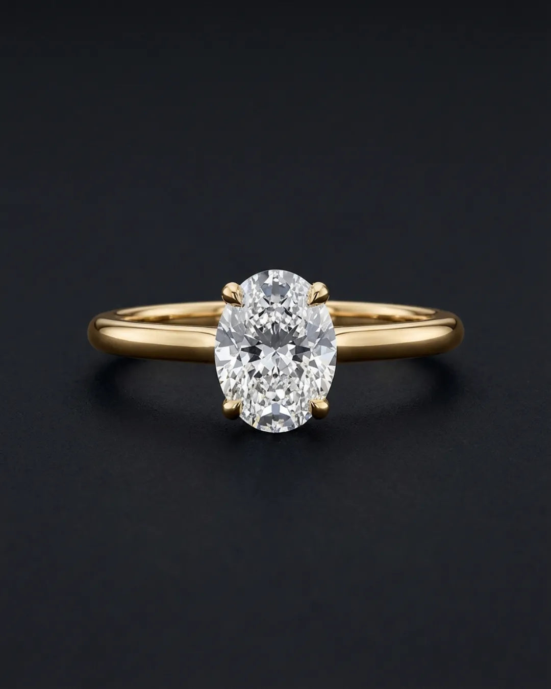 Ovalslipet lab-diamant forlovelsesring i gult gull med fire klør og slank skinne, fotografert mot mørk antrasitt bakgrunn.