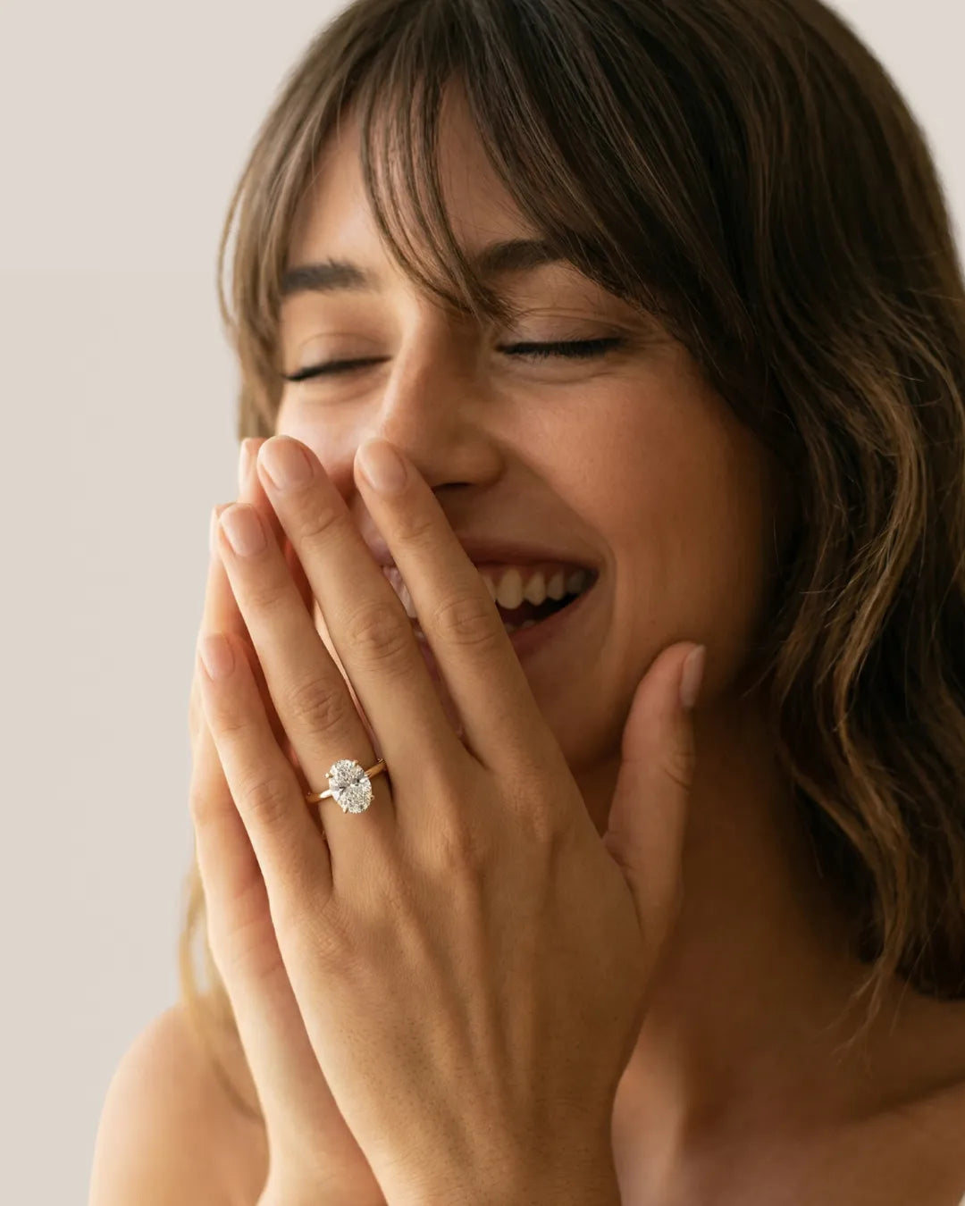 Ung kvinne med brunt hår ler med lukkede øyne og hendene løftet mot ansiktet med ovalslipet lab-diamant forlovelsesring i gult gull på ringfingeren.