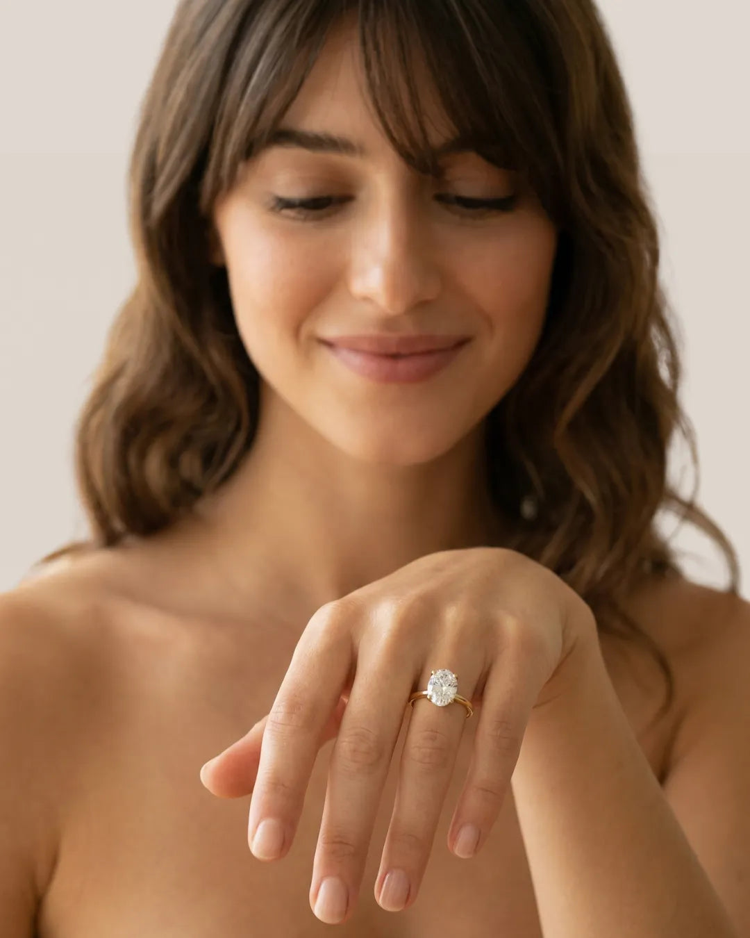 Ung kvinne med brunt bølgete hår ser ned og smiler mot en ovalslipet lab-diamant forlovelsesring i gult gull på ringfingeren.