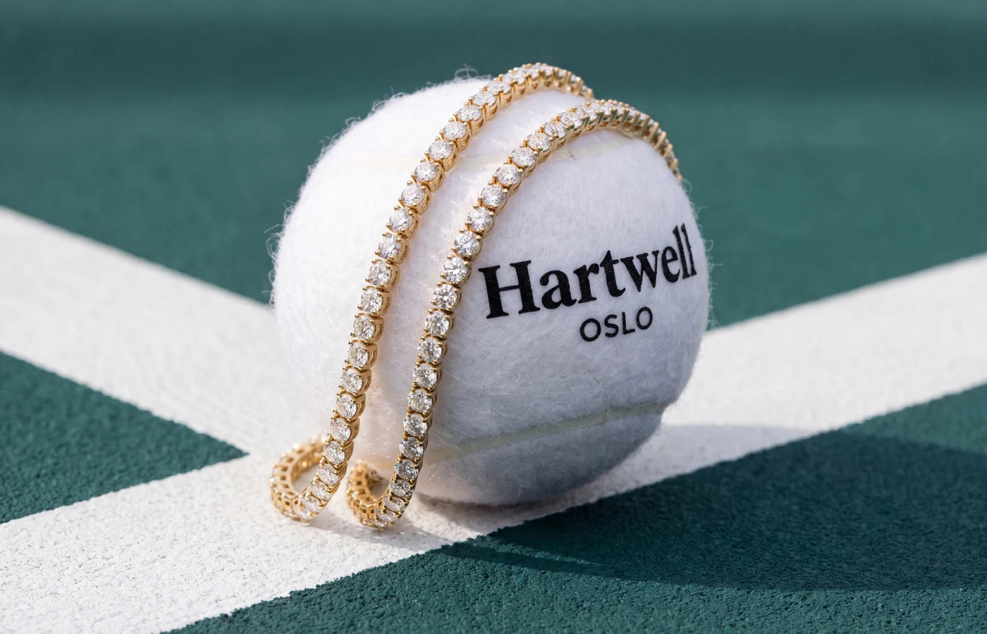 T tennisarmbånd i gult gull ligger over en hvit tennisball med Hartwell oslo  logo på en tennisbane. 